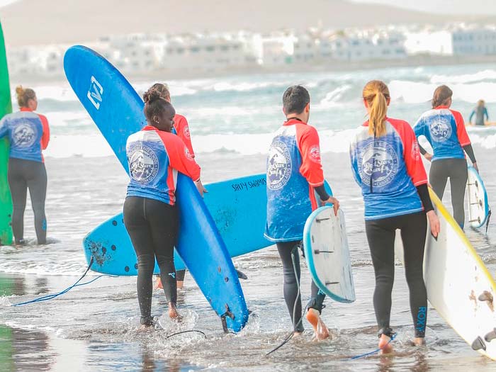Imagen de varias personas en la orilla haciendo surf, cargando con las tablas, todos llevan la misma camiseta. Como si de una escuela se tratara.